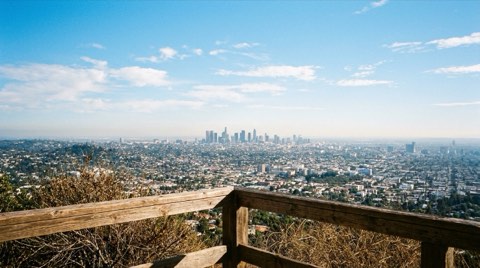 A clear daytime view of the downtown Los Angeles skyline seen from a Hollywood Hills vantage point, with a hillside deck railing and dry brush framing the foreground