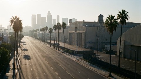 A wide arterial road leading toward distant studio-lot style soundstage buildings in morning commute light.