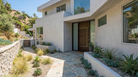 A well-maintained Hollywood Hills hillside home exterior in bright daylight, with a front entry path, clean stucco facade, and manicured landscaping, as seen when arriving for a showing
