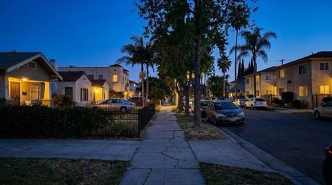 A residential Los Angeles street at blue hour dusk, with apartment buildings and single-family homes showing warm window light against a deepening blue sky.