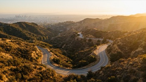 A golden-hour view along Mulholland Drive's winding ridge road in Hollywood Hills, with dry chaparral hillsides and a hazy city basin silhouette visible in the distance