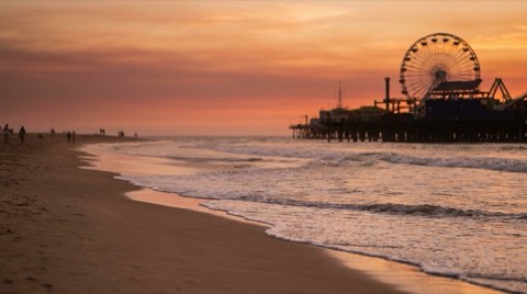 A wide sandy Santa Monica beach at golden hour with the pier and Ferris wheel silhouetted against the sunset